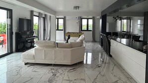 A spacious living area with a large white sofa, a piano, and a kitchen area with a long counter. - Beautiful house with private pool Beaujolais - 35 min from Lyon (Gleizé)