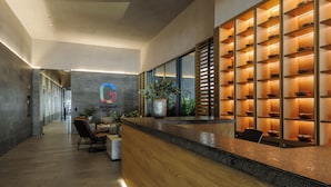A modern reception area with a wooden reception desk, a large potted plant, and a wall-mounted shelf with bowls. - Be grand del Valle by Wynwood House (Mexico City)