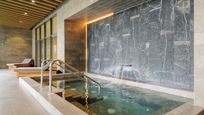 A modern indoor pool with clear water, a stone wall with a waterfall feature, and wooden loungers. - Be grand del Valle by Wynwood House (Mexico City)