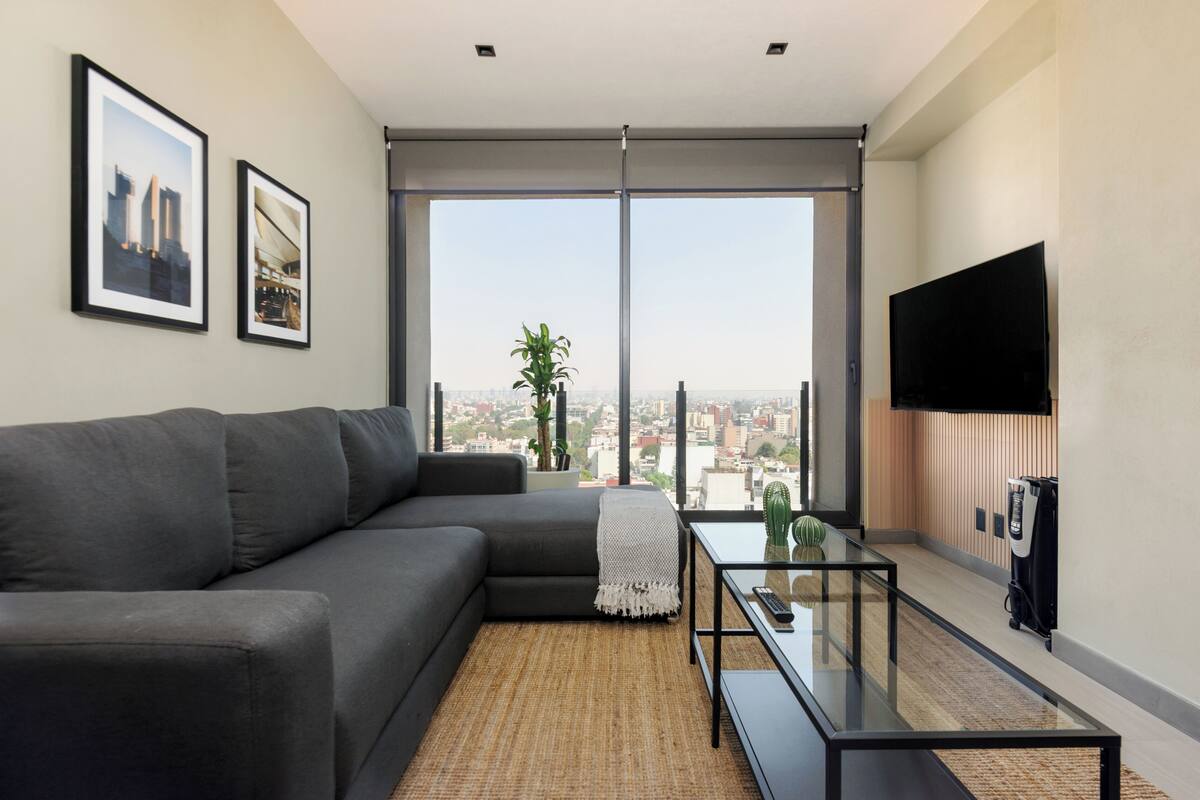 Una sala de estar moderna con un sofá gris, una mesa de centro de vidrio, un televisor de pantalla plana y un amplio ventanal con vistas a la ciudad.