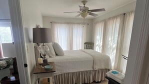 Ein Schlafzimmer mit Bett, Nachttisch, Lampe, Deckenventilator und einem Fenster mit Vorhängen.