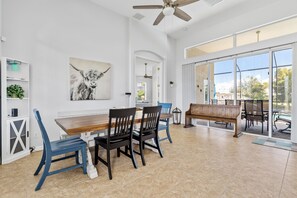 A spacious dining area with a wooden table and chairs, a ceiling fan, and large windows offering a view of the outdoors. - Serene Place 2-Cape Coral (Cape Coral)