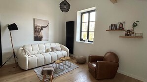 A living room with a sofa, a chair, a floor lamp, a radiator, a window, and a magazine on a shelf. - Le Nid - Domaine de la Tour - New apartment for 6 people (Fleury)