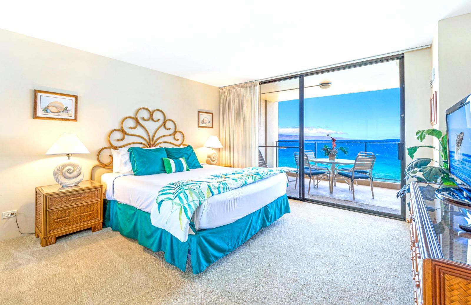 Habitación de hotel con cama, mesita de noche, televisor y balcón con vista al océano.