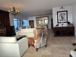 A spacious living room with a white sofa, a patterned ottoman, a wooden cabinet, and a large mirror.