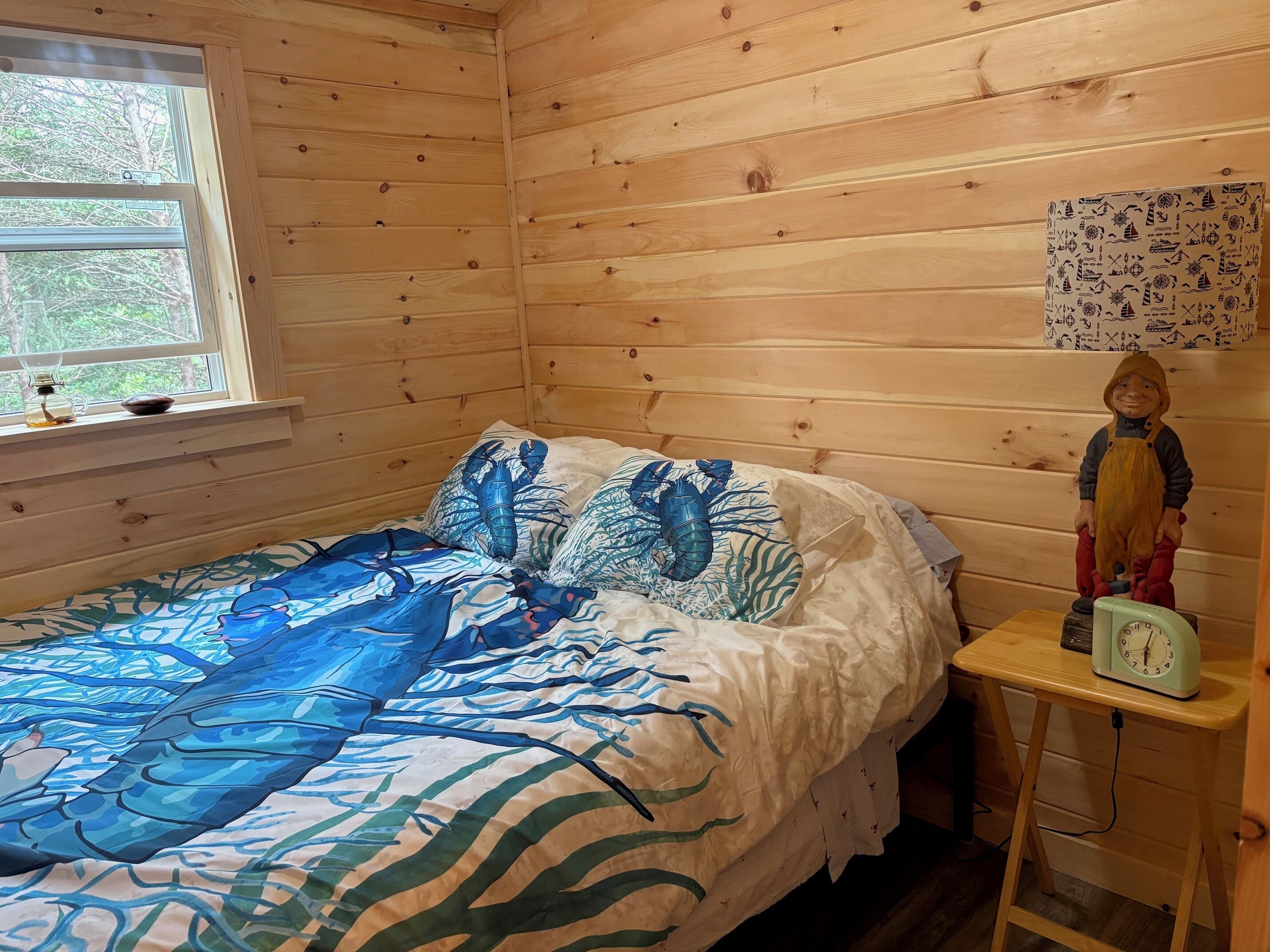 A wooden cabin bedroom with a bed, a nightstand, a clock, and a window with a view of trees.
