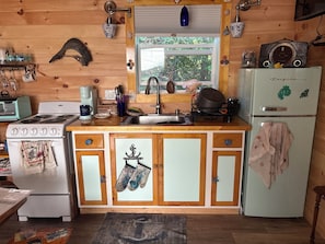 A compact kitchen with a stove, sink, and refrigerator, wooden cabinets, and a window with a view of greenery.