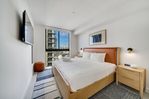 Una habitación de hotel moderna con una cama grande, un televisor de pantalla plana montado en la pared, una mesita de noche con una lámpara y vistas a la ciudad a través de un ventanal amplio.