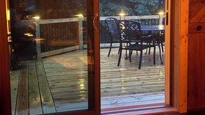 Una terraza de madera con un ventanal de vidrio que da a un área de estar exterior con sillas y una mesa.
