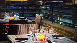 Un restaurante con terraza, mesas dispuestas con vasos y cubiertos, y vistas a edificios al fondo.
