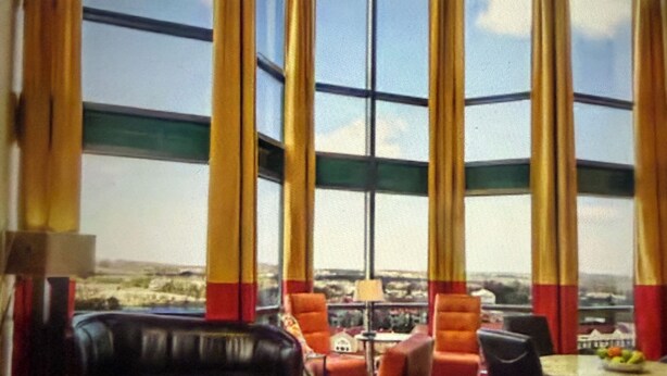 Un amplio salón con un sofá de cuero negro, sillones naranjas, una mesa de centro redonda y una mesa de comedor con sillas. Grandes ventanales ofrecen vistas al exterior.