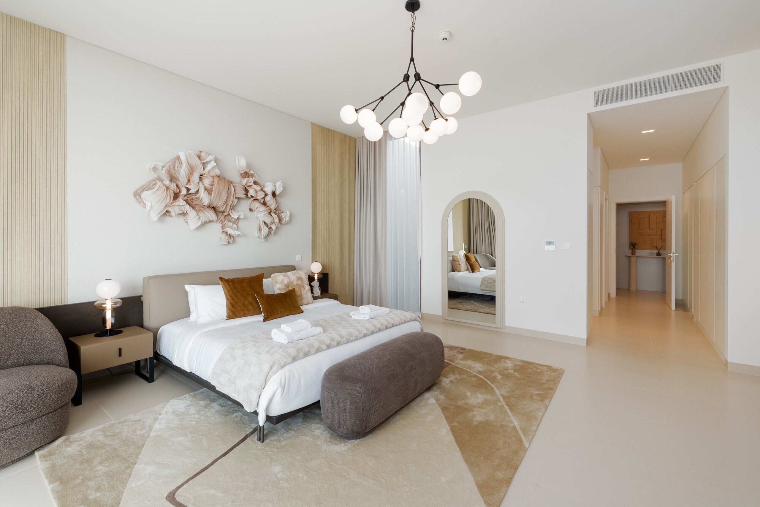 A modern bedroom with a large bed, a bedside table with a lamp, a mirror, and a view into another room.