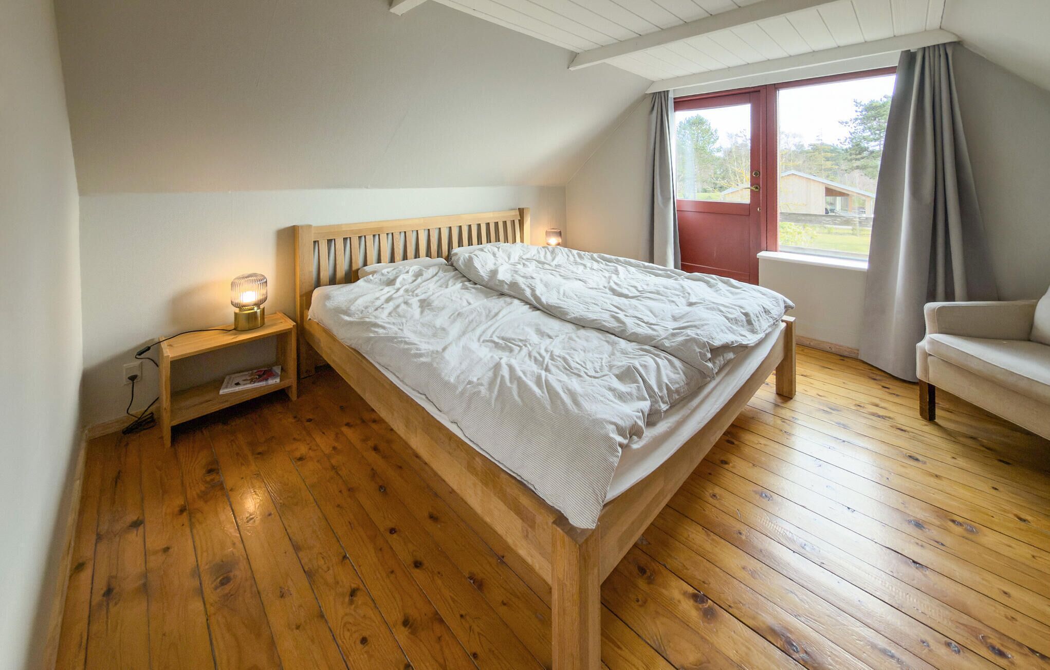 Een slaapkamer met een houten bed, een nachtkastje met een lamp, een raam met gordijnen en uitzicht op de omgeving.