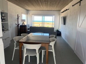 A dining area with a wooden table and white chairs, a sofa, and a large window with a view of the beach.