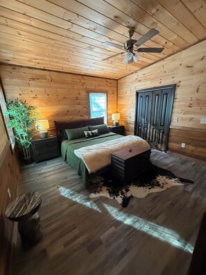 Ein Schlafzimmer einer Holzhütte mit einem Bett, Nachttischen, einem Deckenventilator, einem Fenster und einer Tür.