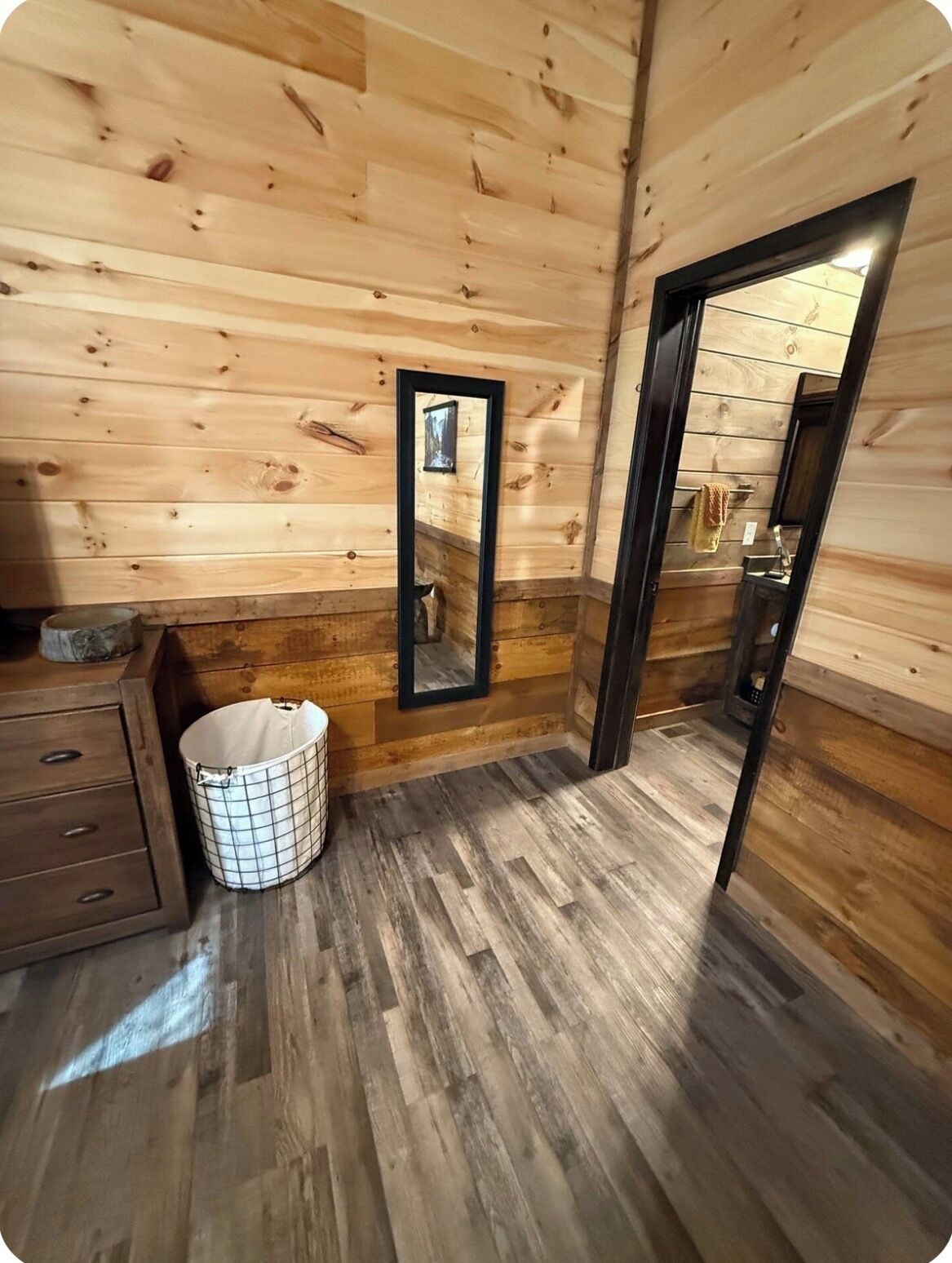 A wooden interior with a wooden floor, a mirror, a small trash bin, and a wooden cabinet.