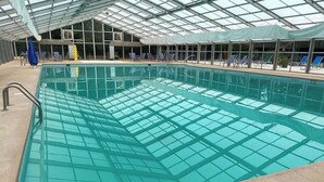 Una piscina interior con paredes de vidrio transparente, sillones de descanso y una zona cubierta.