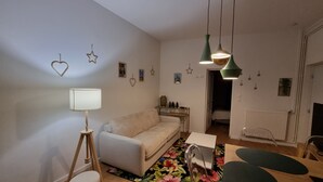 Un salon chaleureux avec un canapé, une petite table, une lampe et un tapis à motifs floraux. Des décorations en forme d’étoile ornent le mur, et une porte donne accès à une autre pièce.