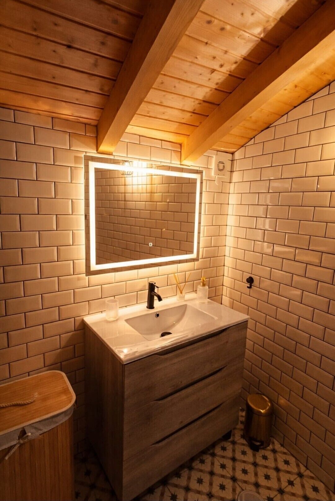 Een badkamer met een houten plafond, een grote spiegel met verlichting, een wit wastafelmeubel en een vloer met een patroon.