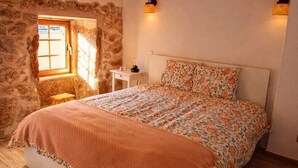 Quarto com teto de madeira, parede de pedra, cama com roupa de cama floral, tapete tecido e uma pequena mesa com uma chaleira.