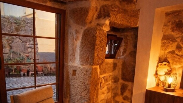 Um quarto aconchegante com parede de pedra, uma cadeira de balanço de madeira, uma poltrona estofada e uma pequena mesa com um candeeiro.