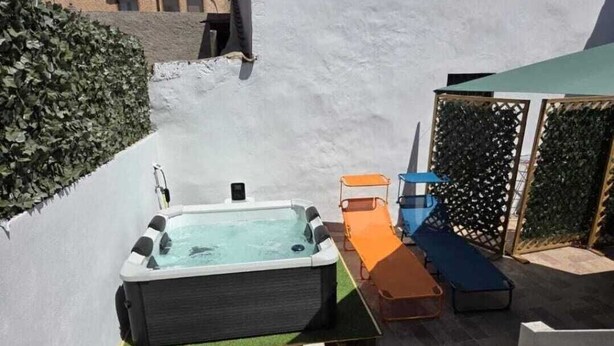 Un jacuzzi con una pequeña piscina, rodeado por un muro blanco y un seto verde. Hay dos sillones de relax, uno naranja y otro azul, y una valla de madera al fondo.