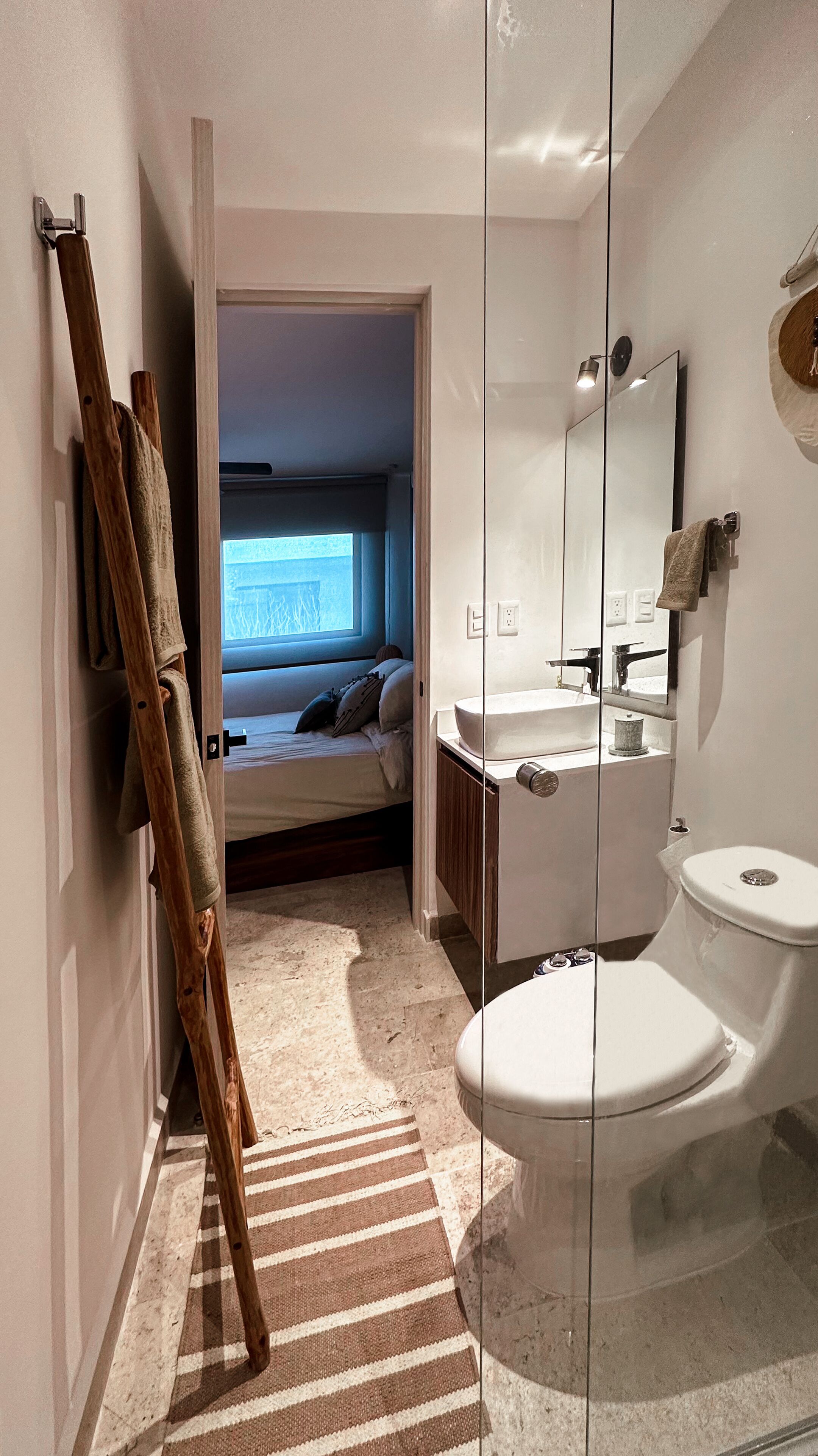 A modern bathroom with a glass-enclosed shower, a white sink, and a toilet. There is a wooden ladder leaning against the wall and a striped rug on the floor.
