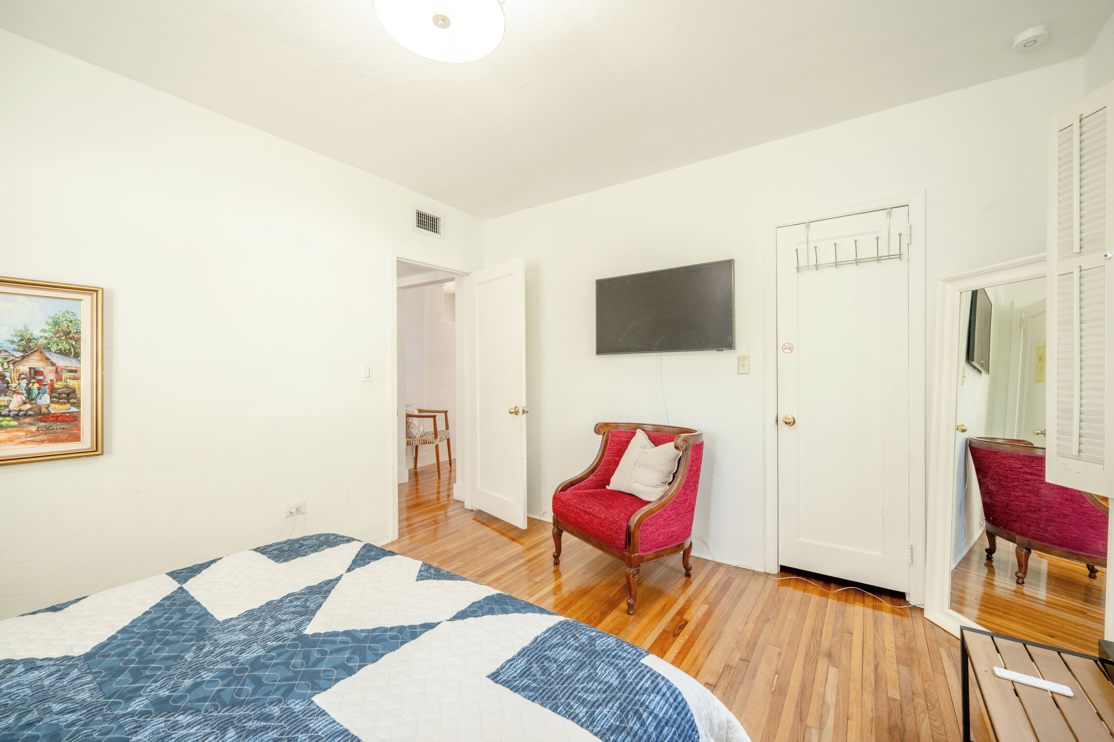 A bedroom with a bed, a red armchair, a TV mounted on the wall, and a door leading to another room.