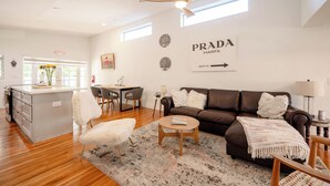 Een moderne woonkamer met een bruine leren bank, een witte stoel en een salontafel. Er is een keuken met een wit kookeiland en een eettafel met stoelen. De kamer heeft een plafondventilator en een bord op de muur waarop staat: PRADA MARFA 1937 MI.