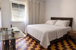 A bedroom with a bed, a glass table, a wooden nightstand, and a window with curtains. - Apto Pet Friendly com Ar condicionado no Centro (Bagé)
