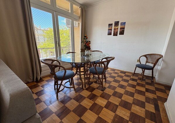 A dining area with a glass table surrounded by wicker chairs, a checkered wooden floor, and a view through large windows.