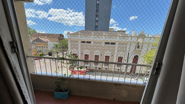 A balcony with a view of a building with a tall tower, a potted plant, and a red vehicle on the street.