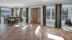 A spacious room with a dining area, a large wooden door with a decorative glass panel, and large windows with curtains. - Pond View Retreat | Bright & Spacious 3BR (West Columbia)