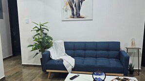 Una sala de estar con un sofá azul, una mesa de centro blanca y un tapete con estampado geométrico, un cuadro de elefantes en la pared y una planta en maceta.