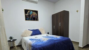 Un dormitorio con cama, aire acondicionado, armario, una mesita con una planta y un cuadro en la pared.