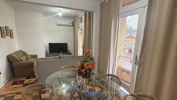Un salon avec une table à manger dont le plateau est en verre, entourée de chaises en osier, d’un canapé et d’un téléviseur placé au fond de la pièce.