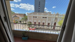 Un balcon d’où l’on peut admirer un immeuble doté d’une haute tour, un pot de fleurs et un véhicule rouge dans la rue.