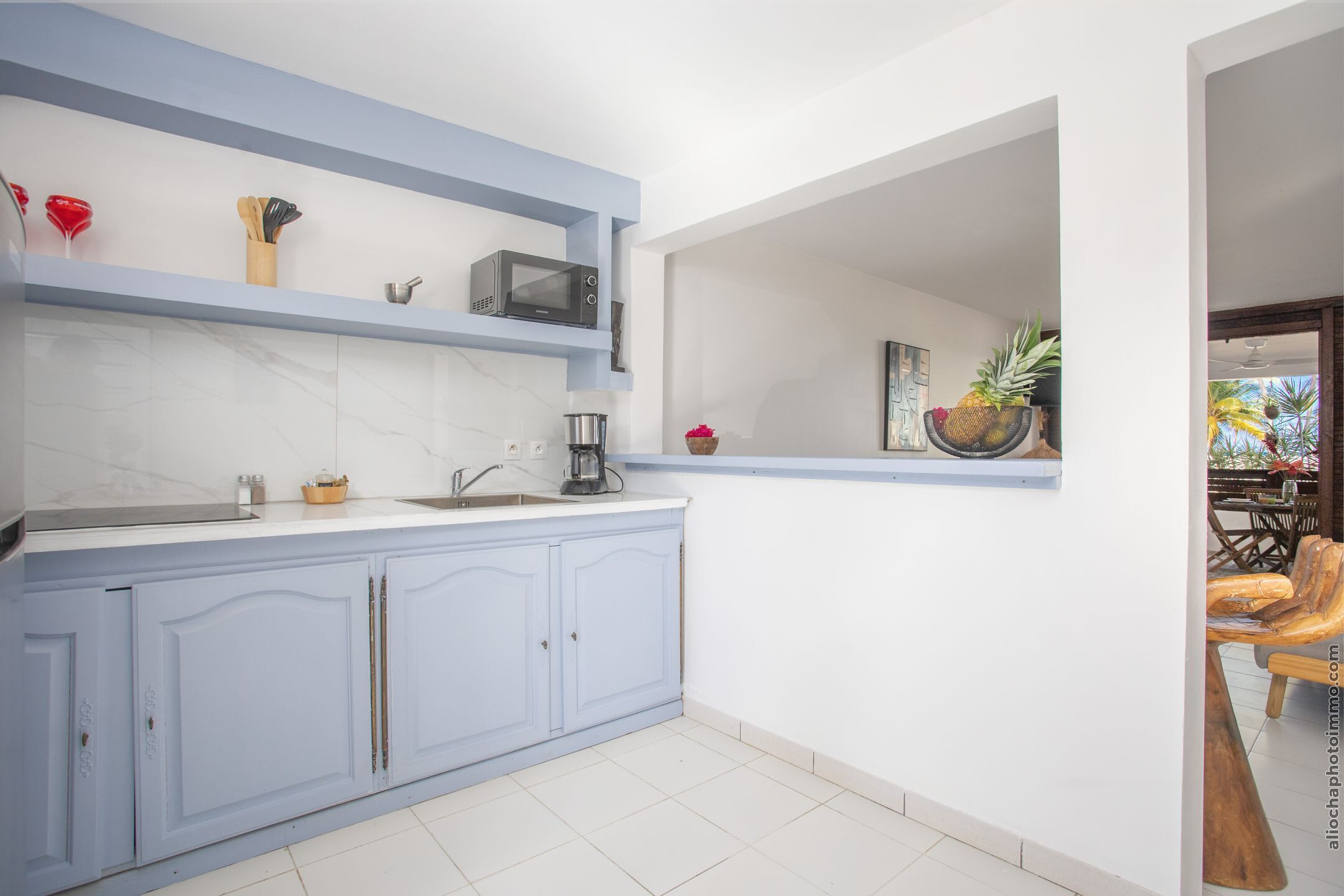 A modern kitchen with light blue cabinets, a white countertop, and a microwave. There is a coffee maker and a bowl of fruit on the counter.