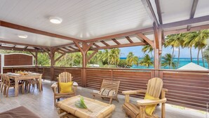 A covered outdoor seating area with wooden furniture, a dining table, and a view of the ocean. - 3-Bedroom Duplex Apartment – Beachfront (La Trinité)