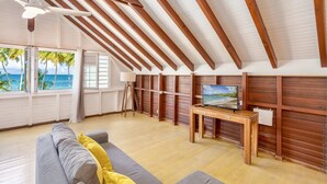 A spacious room with a wooden ceiling, a large window with a view of palm trees and the ocean, a gray sofa with yellow cushions, and a wooden table with a television. - 3-Bedroom Duplex Apartment – Beachfront (La Trinité)
