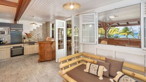 A modern kitchen with a built-in oven, a wooden dining table, and a large window overlooking a tropical beach. - 3-Bedroom Duplex Apartment – Beachfront (La Trinité)