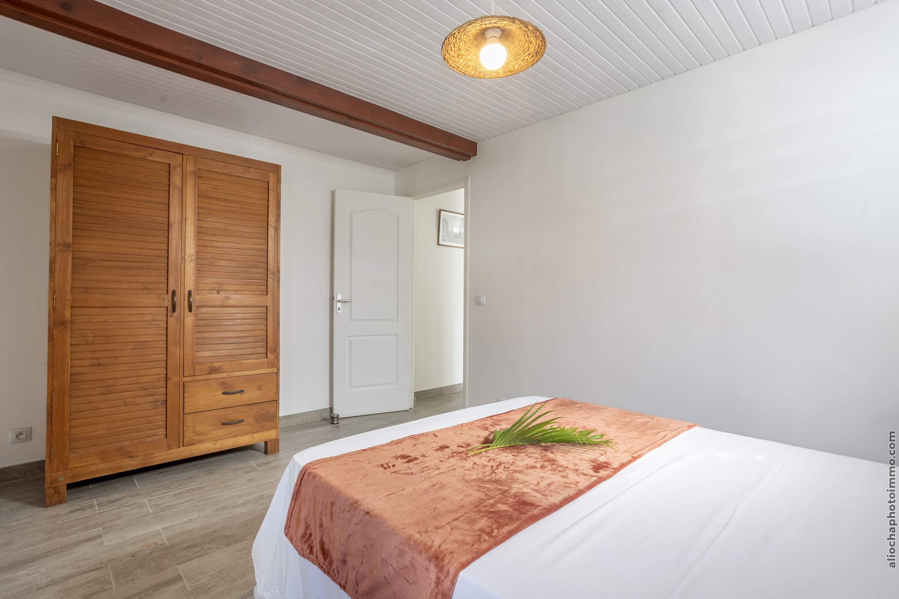 A bedroom with a large wooden wardrobe, a bed with a brown bedspread, and a woven ceiling light.
