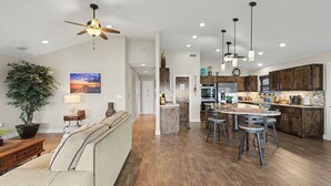 A spacious living area with a sofa, a coffee table, and a large potted plant. There is a kitchen with wooden cabinets and a bar area with stools. - GetawaY at Mi Casita (Rockport)