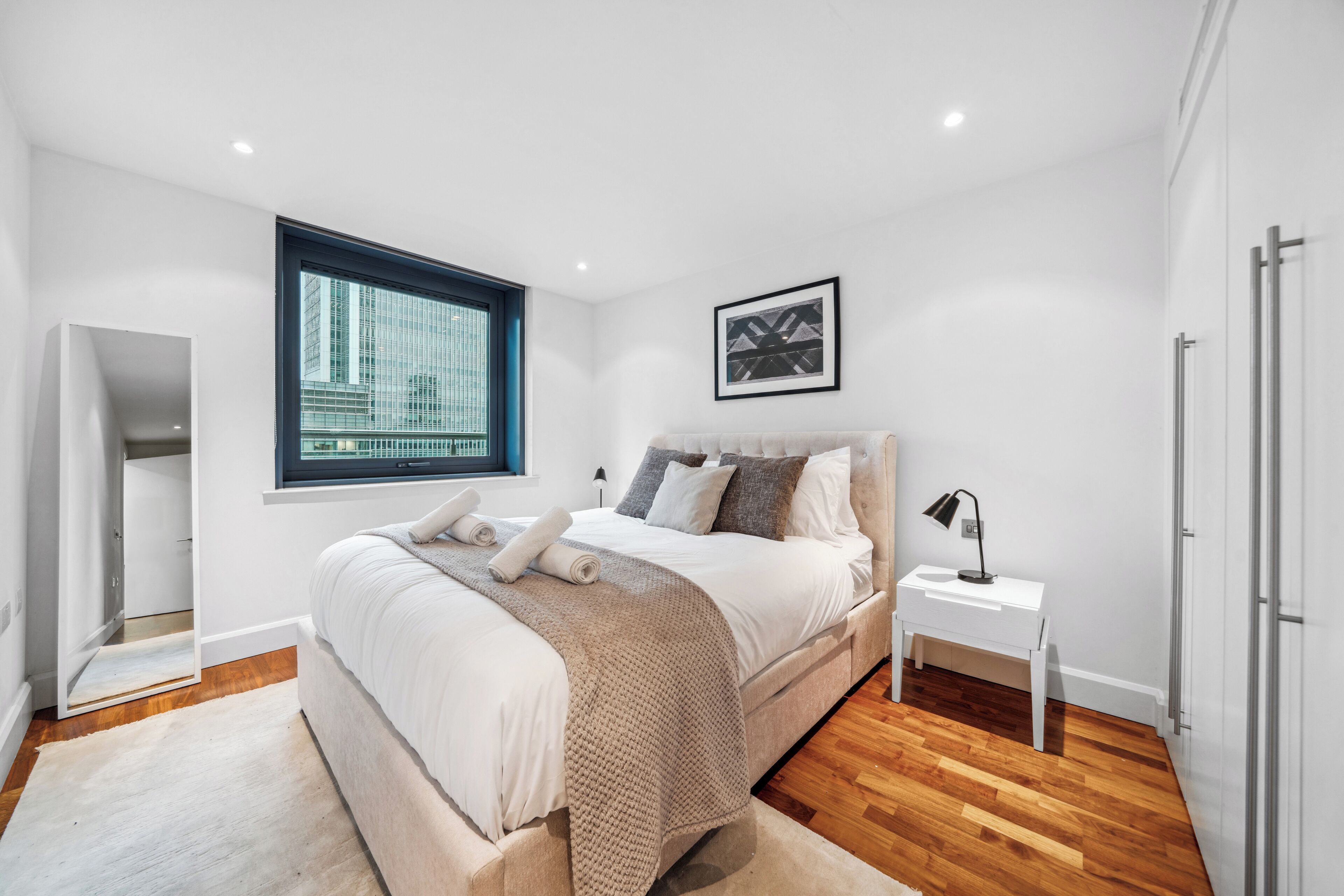 A modern bedroom with a large bed, a bedside table, a window with a city view, and a framed picture on the wall.