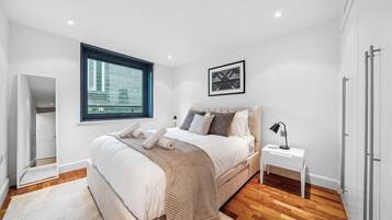 A modern bedroom with a large bed, a bedside table, a window with a city view, and a framed picture on the wall.
