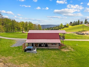 A two-story building with a red roof, a covered porch, and a hot tub in the front yard. - Rustic Barn Retreat w Hot Tub, Game Room & FirePit (Wardensville)