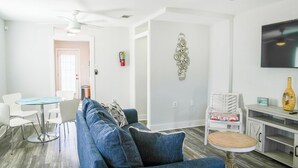 A modern living room with a blue sofa, a round coffee table, and a white dining table with chairs. There is a white wall with a decorative metal piece and a mounted television. - The Southern Porch Cottage Near NOLA & Coast (Slidell)