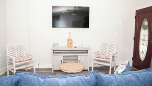 A living room with a TV mounted on the wall, two white chairs with colorful cushions, a small wooden table, and a blue couch. - The Southern Porch Cottage Near NOLA & Coast (Slidell)