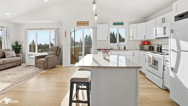 A spacious living room with a sofa and a chair, a kitchen with white cabinets and appliances, and a large window offering a view of trees. - Charming 3-bedroom house in Homer perfect for your getaway (Homer)