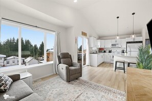 A modern kitchen with white cabinets, a dining area with a wooden table, and a large window offering a view of houses and trees.